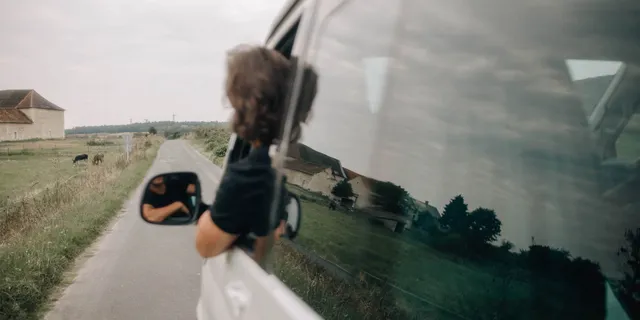 A man leans out of the open window of a camper and looks at the road that the camper travels long.