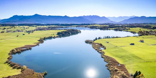 Riegsee camping: A top view of Lake Riegsee in the Bavarian Alpine foothills, surrounded by lush green meadows with a mountain panorama in the sunshine.