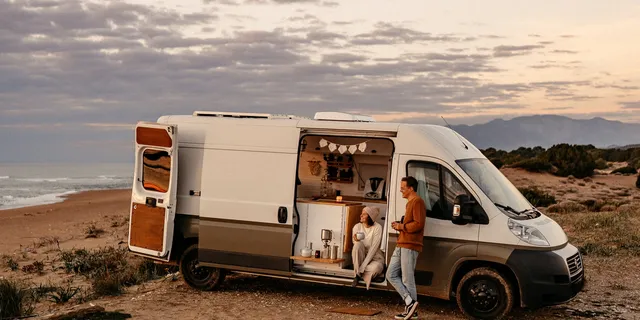 A woman sitting in a parked CamperBoys van and a man outside leaning against the van. They are both drinking something and  looking happily into the same direction.
