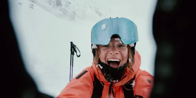 A woman is laughing at the camera, wearing a ski outfit, with snow visible behind her.