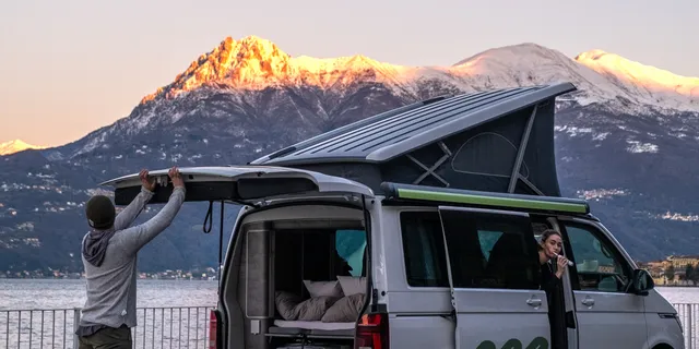 A young man just closes the trunk of his off camper, on the passenger's seat a young woman brushes teeth. Behind there is a lake and a mountain with snow.