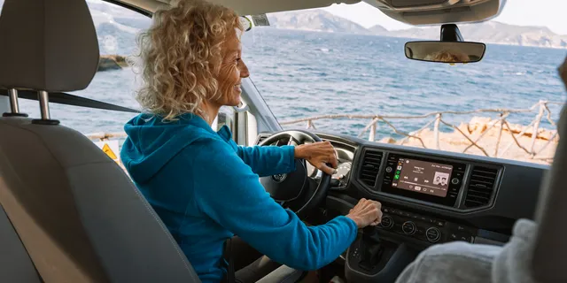 Eine ältere Frau mit blauem Pulli sitzt in der Fahrerkabine ihres Campers und macht das Radio an.