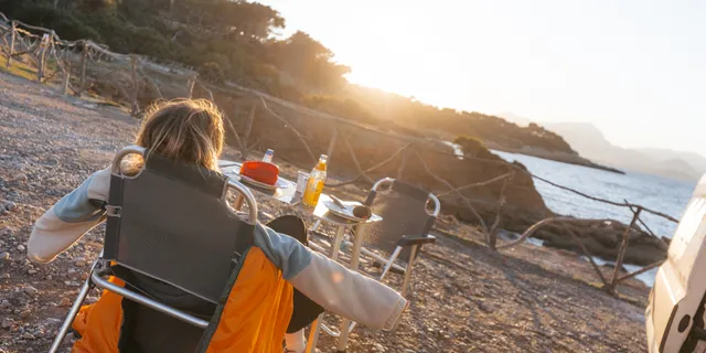 Mit Camping Tipps können Camper-Neulinge entspannt verreisen: Wie die junge Frau, die im Campingstuhl vor einem Tisch in der Sonne aufs Meer schaut und die Arme zur Seite streckt.