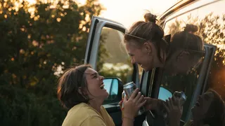 Ein lesbisches Pärchen lacht sich aus dem Fenster eines Campers an und hält eine Getränkedose in der Hand.