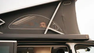 Ein junger Mann schaut beim Aufbauen seines Camper Aufstelldachs aus dem Fenster nach draußen.