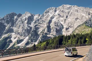 Ein weißer Campervan mit Fahrrädern fährt eine Straße entlang, im Hintergrund sieht man eine Bergkette, Wälder und blauen Himmel