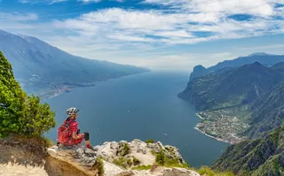 Person in mountain biking equipment sits on rock and overlooks Lake Garda from above