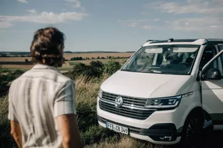 A man looks at his Off Camper VW Bus, which parks on the roadside next to a field