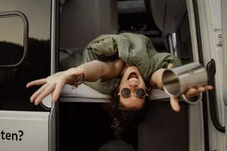 A woman stretches out her arms laughingly from the open rear door and holds a camping cup in her hand.