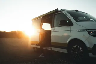A VW California Van in the sunshine. A woman sits in the open sliding door and ties the shoes.