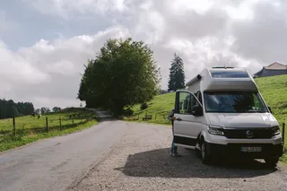 A Grand California Van is parked in the mountains. A man opens the sliding door. On a pasture next to grass cows.