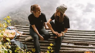 Two young women sit on a ridge during their Riegsee camping to take a break while walking and laugh at
