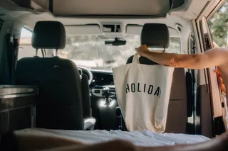 Inside a VW California Ocean Van, a girl hangs a linen bag with the inscription 'Holiday' over the passenger seat headrest.