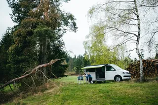 She sits under awning in front of a VW Ocean in the forest.