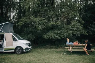 At the camping in Estonia there is a girl on a picnic table, her father sits on the bank. A VW California Ocean Van is also parked.