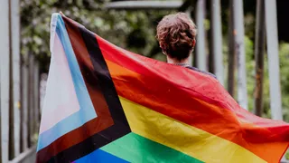 A person photographed from behind, holds the colorful rainbow flag of the LGBTQIA+ community in his hands.