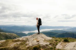 A man with walking backpack on the back stands up on a mountain, in the background you see mountains and water.