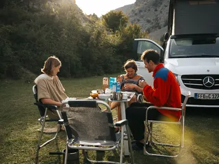 A group of friends sits around a camping table during breakfast at Camping im Münstertal. In the background a Mercedes Marco Polo camper.