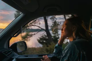 Eine Frau sitzt auf dem Beifahrersitz eines fahrenden Campers beim Überwintern in Portugal. Man sieht draußen den Sonnenuntergang.