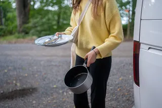A young woman in yellow sweater wears dirty plates, cutlery and a pan for washing - with clean water tank in the camper.