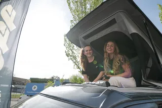 Two young women sit laughing at the camper canopy and enjoy the sun before they go on holiday with their off camper.