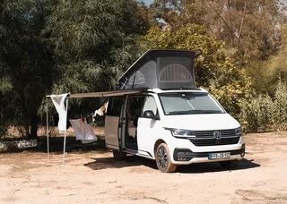 Ein Camper steht dicht bei einem Wald mit ausgefahrener Markise.