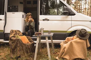A Grand California Van is parked in an autumn forest. A woman sits in the open sliding door with a warm drink, a camping table and chairs are built in front of the van.