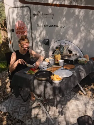 A woman is sitting at a camping table set with breakfast in front of the door of a white Vanever Corsica model motorhome.