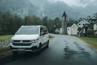 A camper is on a street. A woman leans out of the window and looks at the mountains.