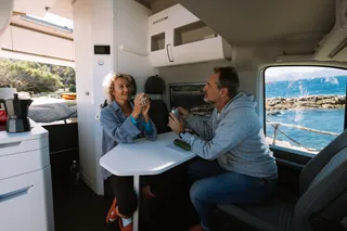 Electricity in the camper is the A and O of a camper trip: A couple sits in the camper's living room by the sea, entertains and drinks coffee.