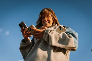 A young woman holds three cell phones at the same time in her hands and laughs against the bright blue sky in the background.